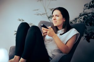 A woman sitting in a chair holds a remote control squinting at tv, to show needing eye exam with Eye Doctor Near Winfield.