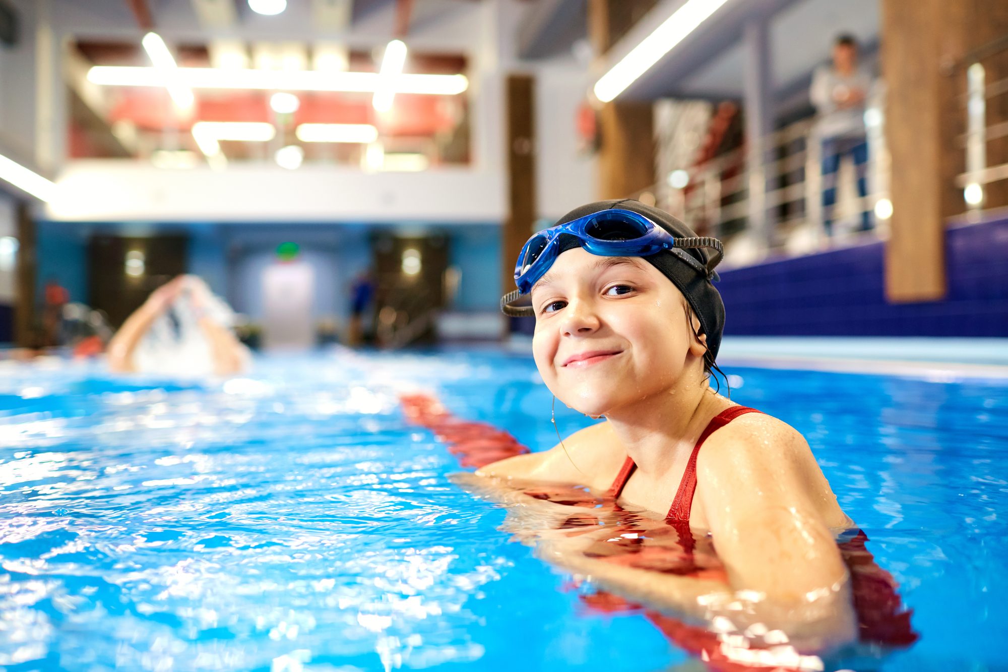 Girl child swimmer in a red bathing suit on a background of the pool inside the room with glasses and a cap, after seeing her Crown Point Emergency Eye Doctor.