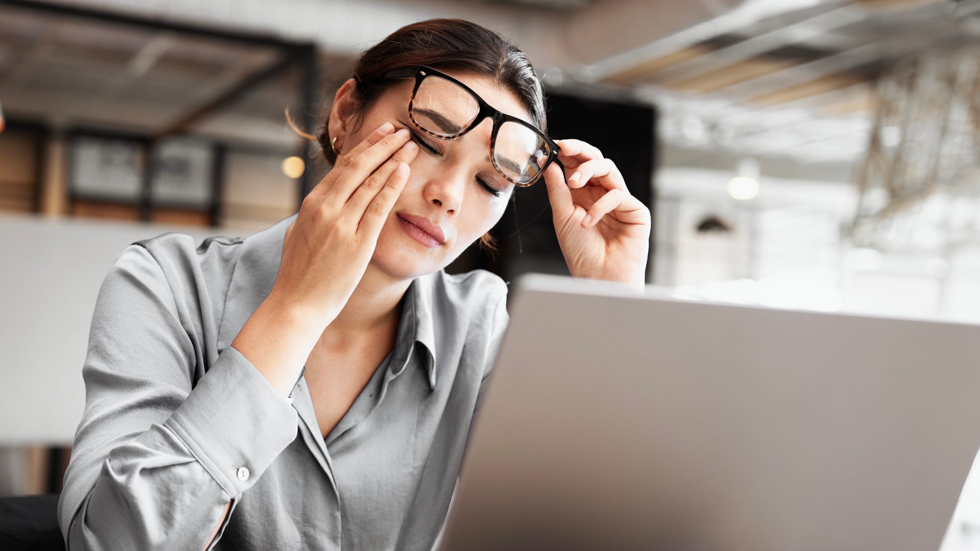 A woman sits at her desk in front of a laptop, removing her glasses and rubbing her eyes, appearing tired—signs that she may be due for a Crown Point Eye Exam to address eye strain in the office.