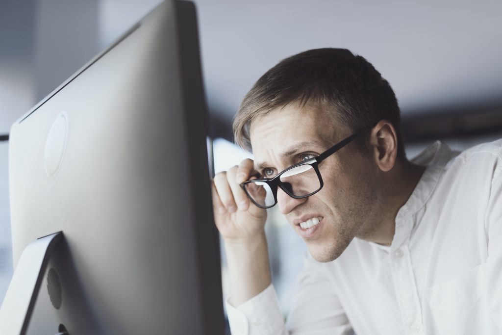 Man squints at computer screen looking over his glasses, to check eyesight make appointment with St. John Routine Eye Exam .