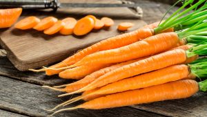 A bunch of fresh carrots with green tops sits on a wooden surface, as if ready for a Lowell eye doctor to recommend them for healthy vision; nearby, sliced carrots and a knife rest on a cutting board in the background.