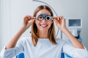 A woman smiles while holding a phoropter to her eyes during her St. John routine eye exam in a bright optometry clinic.