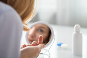 Person holding a contact lens on their fingertip in front of a mirror, with a contact lens case and solution bottle on the table—preparing for the day after their Cedar Lake contact lens exam.