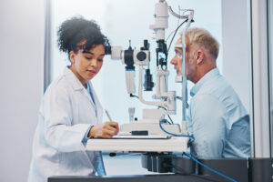 An eye doctor conducts a St. John vision exam on a male patient using a slit lamp while taking notes in a medical office.
