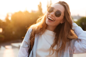 Woman with long hair wearing sunglasses and a white shirt smiles while standing outdoors in sunlight, with one hand touching her hair—showcasing the confidence inspired by St. John Eye Care Center.