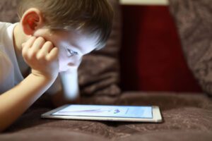 A young boy lies on a couch, resting his chin on his hands and looking intently at a tablet placed in front of him—reminding us of the importance of Lowell children's eye exams.