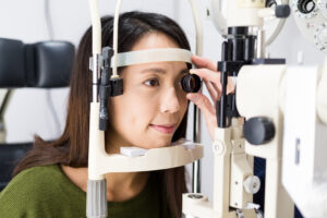 A woman undergoing a Schererville comprehensive eye exam, sitting at a slit lamp while an optometrist examines her eye with a lens.