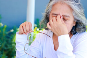 Older woman with gray hair holds her eyeglasses in one hand and pinches the bridge of her nose with the other, appearing tired or stressed while sitting outdoors—an image that highlights the importance of regular Beecher diabetic eye exams.